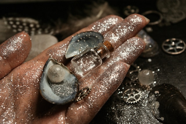 A hand covered in silver glitter holds a shell with a pearl in it.