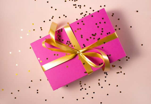 A pink gift box wrapped in gold ribbon and sprinkled with gold confetti.
