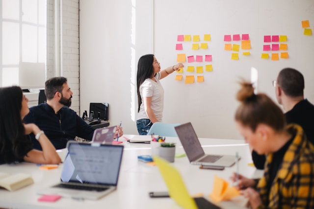A woman stands in front of her team and presents a marketing strategy using post-its on a whiteboard.