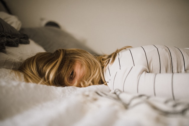 A woman lies on her stomach in bed with one eye peeking out of her hair.