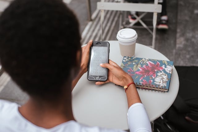 A woman uses her phone while sitting at a table with a phone and a cup of coffee.