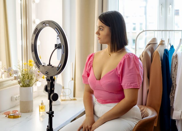 A woman in a pink top sits in front of a ring light.
