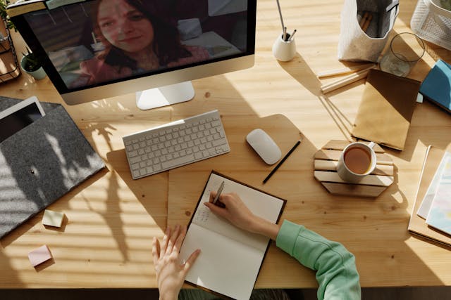 A person writes at a desk and watches a video on a desktop.