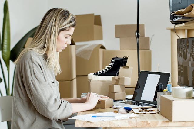 A woman surrounded by boxes types on her laptop.