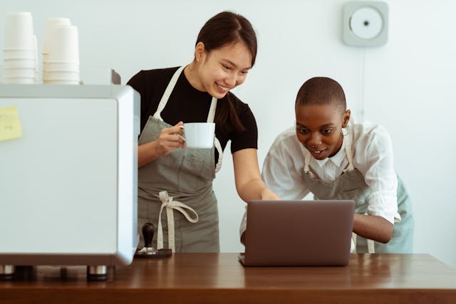 Two women in aprons look at a laptop screen.