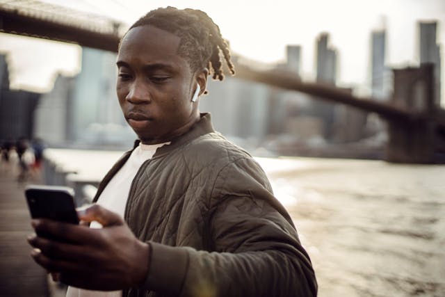A man with AirPods stands on a bridge and scrolls on his phone.
