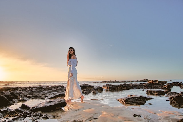 A woman in a long, white skirt and loose, white blouse stands on some rocks by the water.