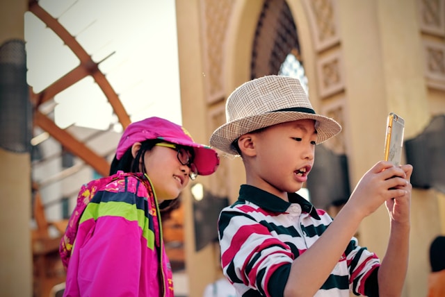 A young boy takes photos with his phone while a young girl stands behind him.