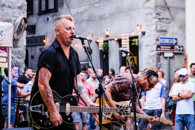 An audience gathers around two male buskers playing guitars and singing out on the street.