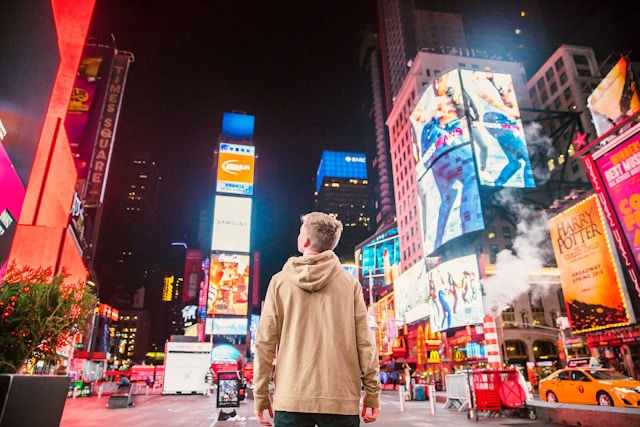 A person looks up at digital billboards looming high on the side of buildings on a busy street at night.