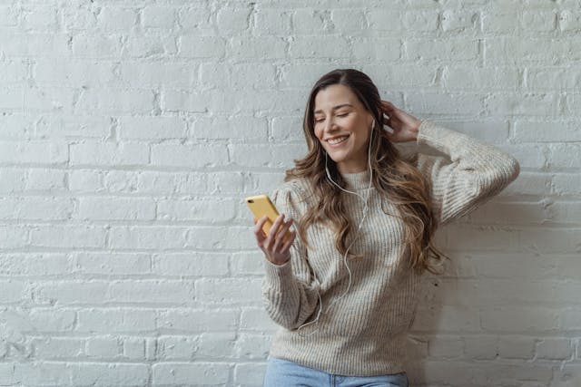 A woman smiles as she listens to her earphones connected to her phone.