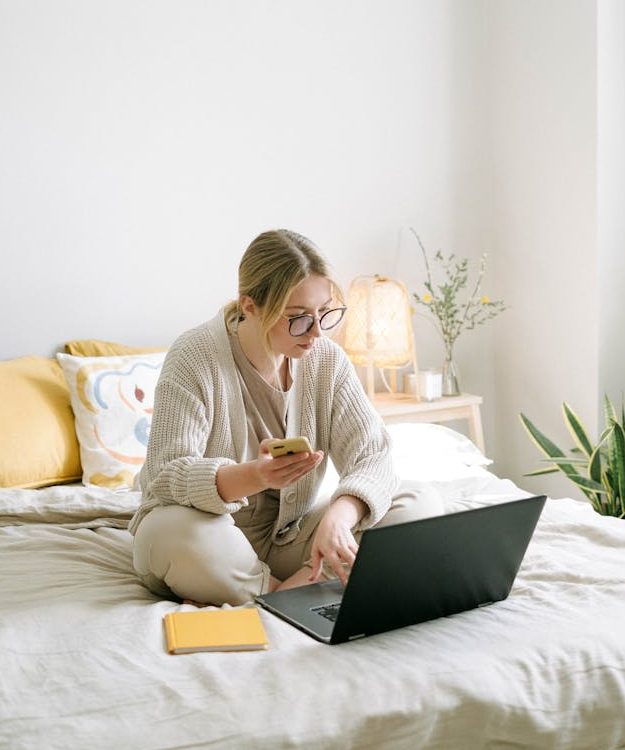 A woman sitting in bed scrolls on her phone and laptop.