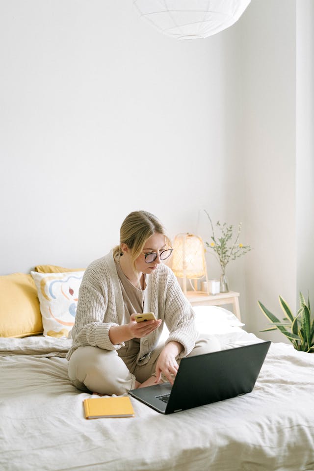 A woman sitting in bed scrolls on her phone and laptop.