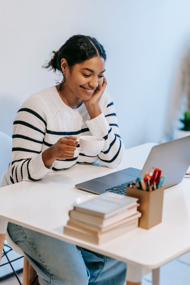 A woman smiles as she looks at her laptop with a cup in her hand.