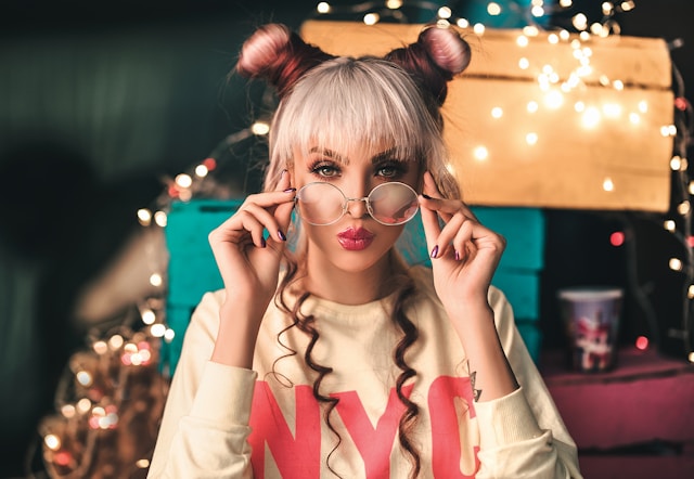A woman with pink and platinum blonde hair is wearing a white sweater, eyeglasses, and pouts for the camera.
