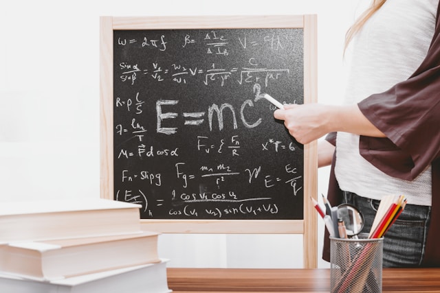 A teacher stands beside a small blackboard with various mathematical formulas on it.
