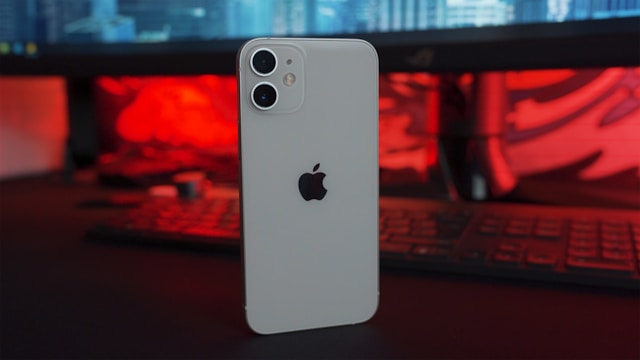 A white iPhone featuring the Apple logo on its back stands on a table in front of a desktop computer.