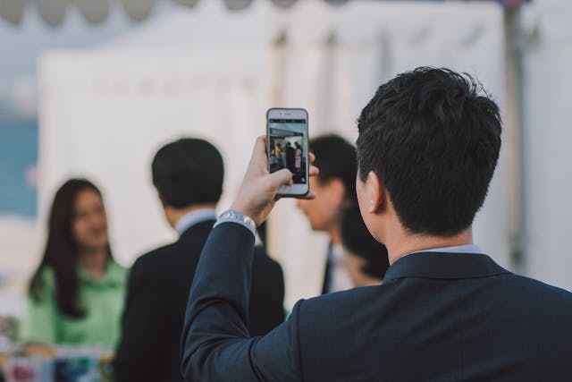 A man wearing a suit takes a photo of a woman speaking.