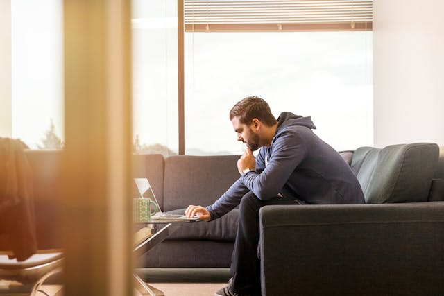A man leans forward on his couch to use his laptop.