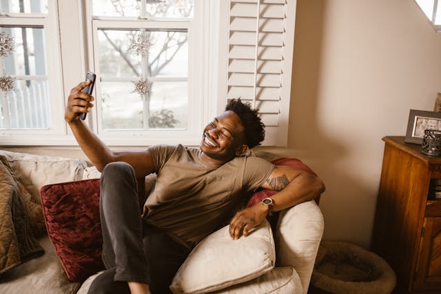 A man grins at his phone screen while he sits on a couch.