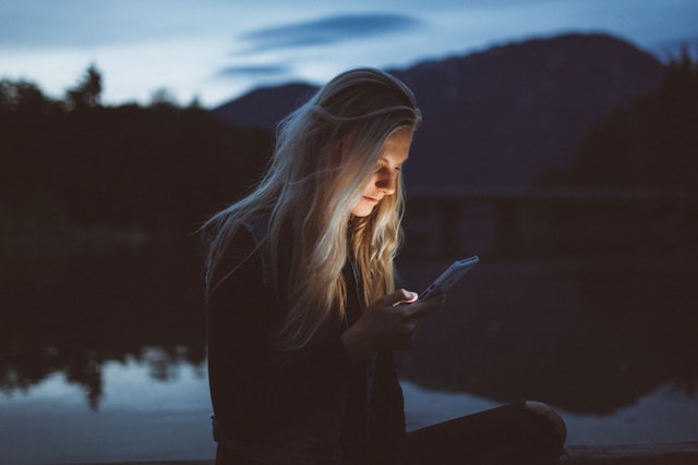 A woman sits by a lake and watches TikTok videos on her phone.
