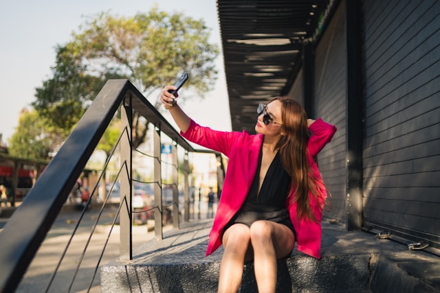 A woman in a pink blazer sits on concrete steps and takes a selfie for her TikTok page.