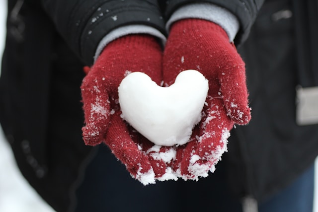 Snow in the shape of a heart in the open hands of a person wearing red gloves.
