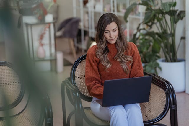 A woman types on her laptop while she sits in a wicker chair.