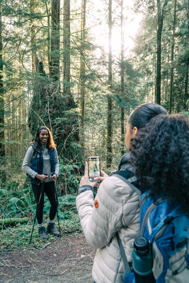 A woman takes a photo of another woman while on a hike in the woods.