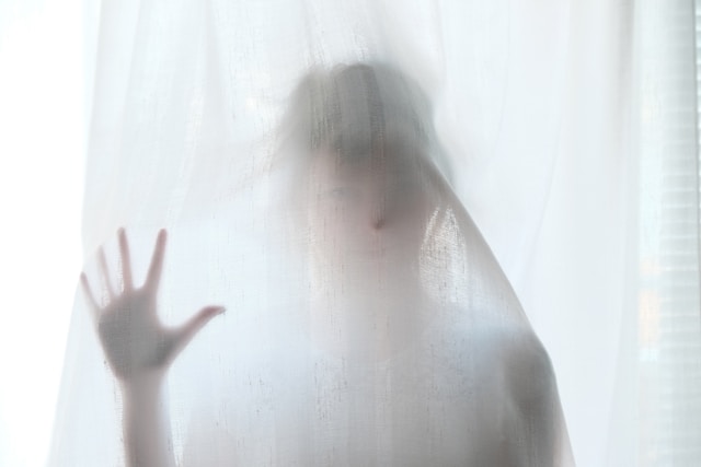 A person hides behind a sheer, white curtain.
