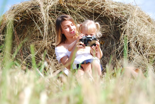 A mom and her daughter sit in front of a hay bale while playing around with an old-fashioned camera.