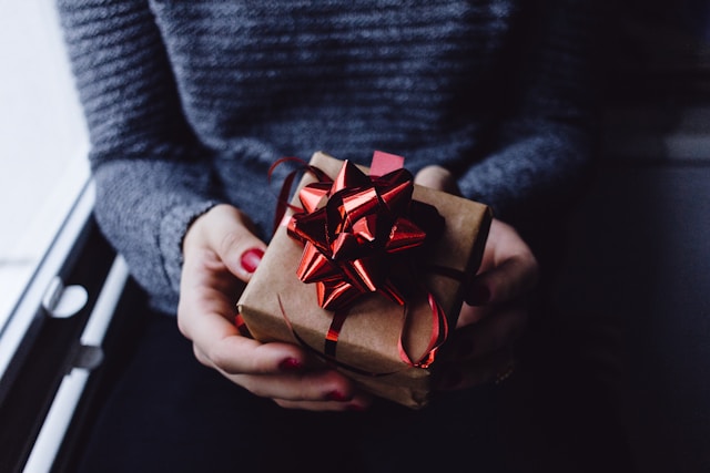 A person holds a gift box wrapped in brown paper and tied with a red ribbon.