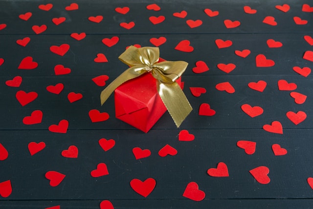 A gift box wrapped in red paper and tied with a gold ribbon sits on top of red paper hearts.