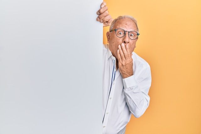 An old man with a shocked expression covers his open mouth with one hand.