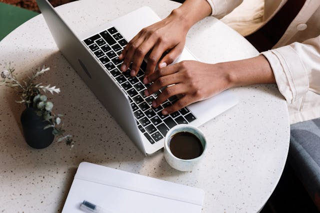 Two hands rest on a laptop beside a cup of coffee.