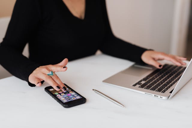 A woman taps on her phone while using a laptop.