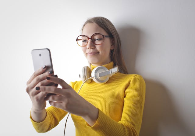 A woman in a yellow turtleneck sweater smiles at her phone.
