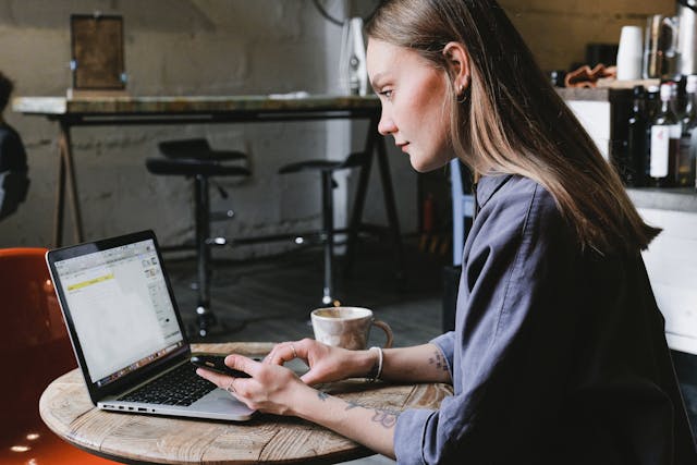 A woman holds her cell phone and types on her laptop at a coffee shop.