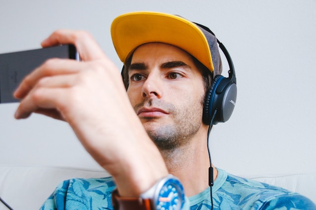 A person wears a cap and headphones while watching TikTok videos on his phone.