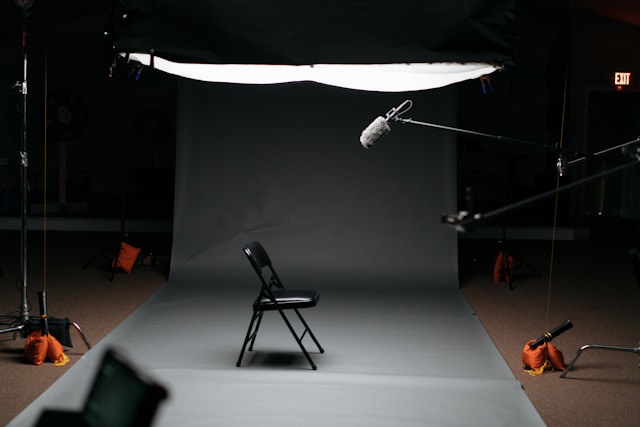 An empty chair sits in front of a gray backdrop, a microphone, and some lighting equipment.