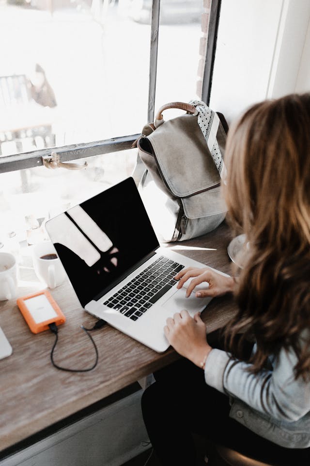 A woman sits at a wooden table to type on her laptop.