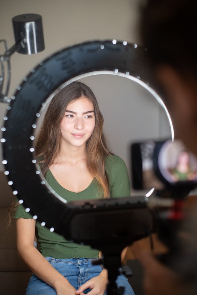 A woman in a green shirt sits in front of a ring light.