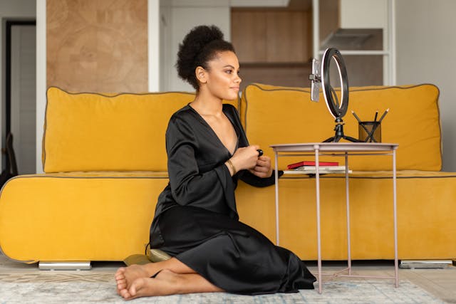 A woman sits by her couch in front of a phone and ring light.