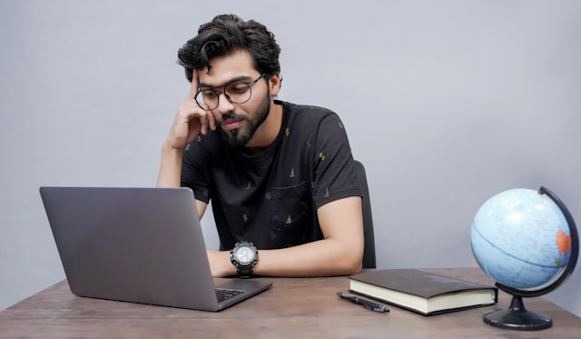 A man sits at a table and stares at his laptop screen, which is next to a book and a globe.