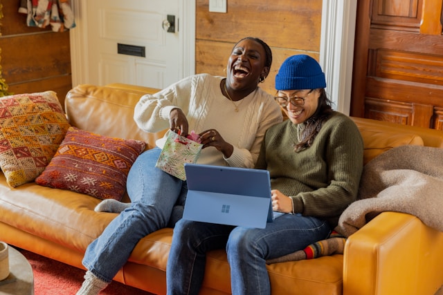 Two friends sit on a couch and share a laugh while watching videos on a laptop.