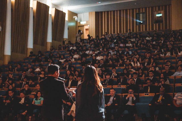 Two speakers stand in front of an audience in an auditorium.